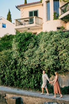 Man and woman walk holding hands along the sidewalk past a high fence overgrown with green ivy. High quality photo