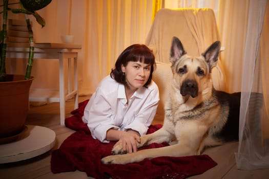 Adult mature woman with big shepherd dog in white shirt. Room with girl and calm cozy evening atmosphere with transparent curtains and soft warm light of lamps. Concept of love for animals, pets