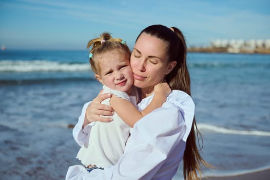 Authentic portrait of young happy affectionate mother gently hugging her adorable little child daughter, walking together on the beach on beautiful warm sunny day. People. Lifestyle. Leisure activity