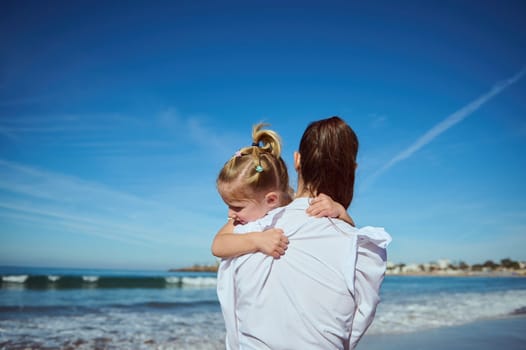 Rear view of a young woman carrying her little daughter, admiring beautiful waves splashing while crashing on the sea shore. Family weekend. Love. Togetherness. People. Healthy active lifestyle