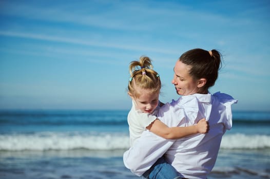 Authentic portrait of a young woman, caring loving mom and her cute little girl daughter, smiling and enjoying happy time together, walking on the beach on warm sunny day. People. Family relationships