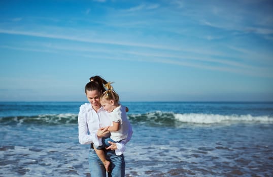 Happy Caucasian smiling young woman, loving affectionate mother carrying little daughter, enjoying a weekend together, walking along a tropical beach. Waves splashing on the background. People. Nature