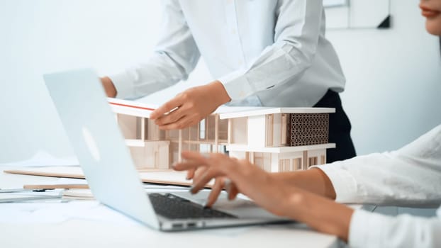 Closeup image of professional male architect hand using ruler to measure house model length while young beautiful caucasian coworker using laptop to analyzed data on meeting table with house model. Immaculate.