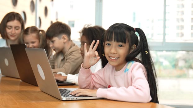 Smart asian girl working on laptop doing ok or okay and looking at camera. Diverse happy children coding or programing engineering code or prompt by using computer in STEM technology class. Erudition.