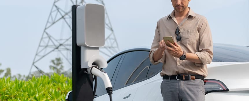 Man pay for electricity with smartphone while recharge EV car battery at charging station connected to electrical lower grid tower for eco friendly car utilization. Panorama Expedient