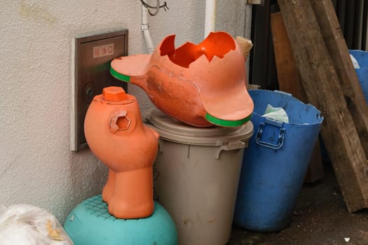 A broken plastic statue pots on a trash bin with other waste containers nearby, depicting waste and recycling.