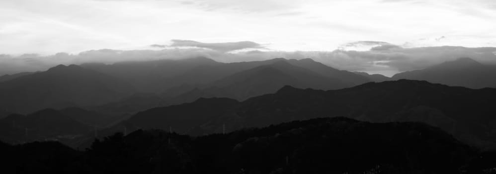 Panoramic black and white image of layered mountain silhouettes under a gradient sky.