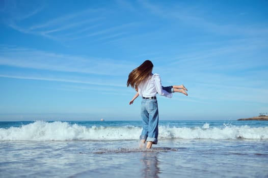 Full length portrait of a mother with long hair, in blue denim, playing with her little child daughter, spinning her around, running barefoot on the waves on Atlantic ocean beach. Happy family pastime