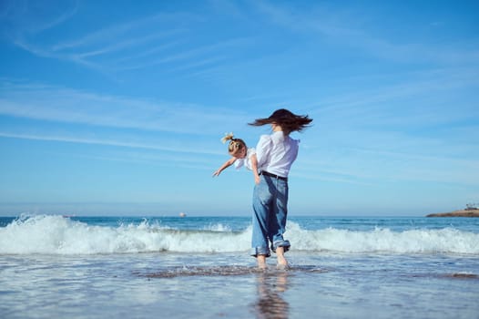 Cheerful young mother playing with her little daughter, running barefoot on the Atlantic beach, jumping on splashing waves crashing on the shore, enjoying together a happy weekend outdoor