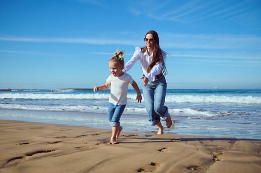 Caucasian cheerful young mother catching her little toddler girl while running barefoot on the beach, enjoying happy time together running on warm water of Atlantic waves splashing on the sea shore