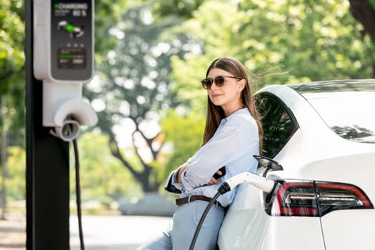 Young woman recharging battery for electric car during road trip travel EV car in natural forest or national park. Eco friendly travel during vacation and holiday. Exalt