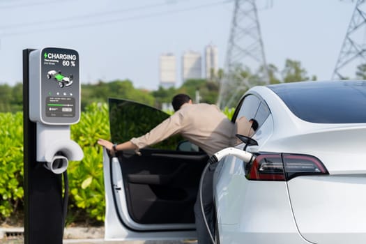 Young man recharge EV car battery at charging station connected to power grid tower electrical industrial facility as electrical industry for eco friendly vehicle utilization. Expedient