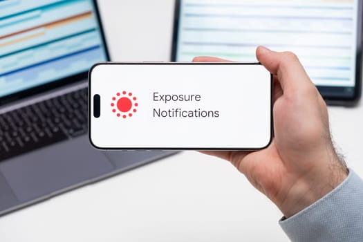 Exposure Notifications application logo on the screen of smart phone in mans hand, laptop and tablet are on the table in the background, December 2023, Prague, Czech Republic.