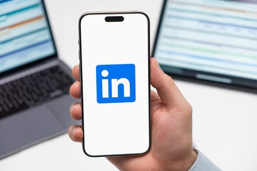 LinkedIn logo of social media application on the screen of smart phone in mans hand, laptop and tablet on the background