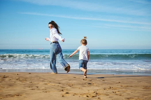 Full length portrait of an authentic family of a young mother and cute daughter, running barefoot on the beach, leaving footsteps on the wet sand while beautiful waves splashing and washing their feet