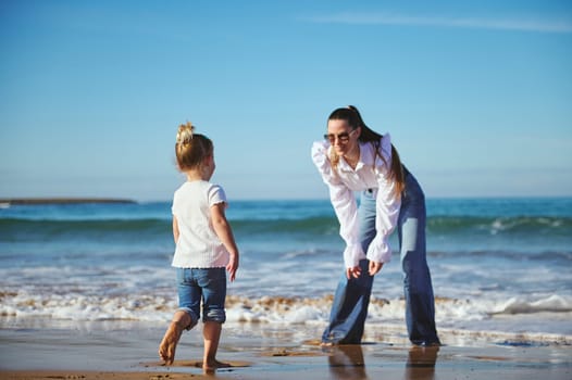 Full length portrait of authentic family of a young mother and her adorable little daughter, standing barefoot on the sandy beach, spending nice weekend on the beach together. Family relationships.