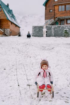 Little smiling girl slides down a snowy hill on a sled against the backdrop of her mom sitting near the house. High quality photo