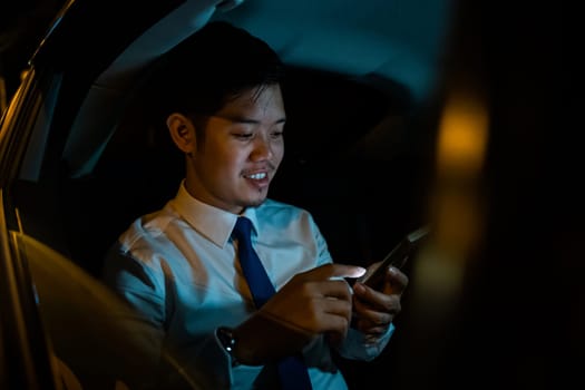 Handsome Asian young man in taxi work on late night sitting back seat and still work with mobile phone in city street, Businessman using smartphone in backseat of car near window at night