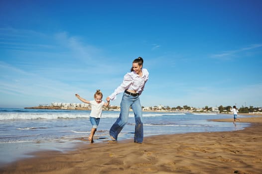 Full length portrait of a Caucasian young pretty woman, loving caring mother and her daughter holding hands and running barefoot on warm water on waves, playing together, spending happy nice time