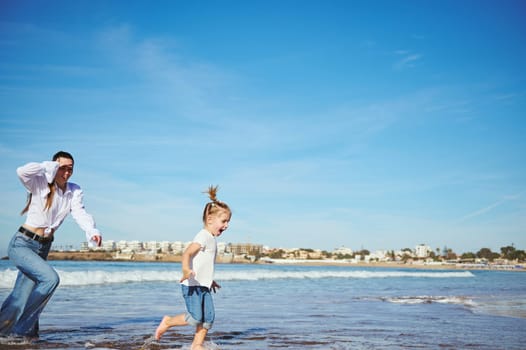 Full length portrait of a young mother and her daughter running barefoot on warm water on waves, playing together, spending happy nice time. Family relationships. People. Lifestyle. Leisure activity