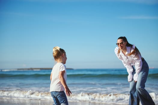 Young happy mother and daughter playing together on the sandy tropical beach on warm sunny day