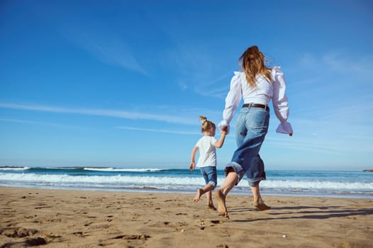 Full length portrait of a Caucasian young mother and her little child girl holding hands while walking barefoot on the sandy beach, dressed together in white shirt and blue denim jeans