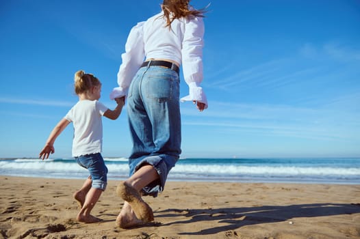 Rear view c of a Caucasian young mother and her little child girl holding hands while walking barefoot on the sandy beach, dressed together in white shirt and blue denim jeans