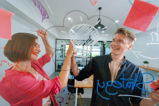 Two smart cooperative business people giving a high five to celebrate the successful start up project while brainstorming marketing idea in front of glass board by using mind map. Immaculate.