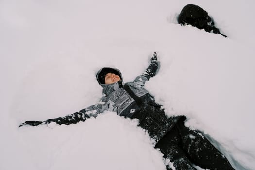 Young smiling woman in a ski suit lies in the snow with her arms outstretched near a black dog. High quality photo