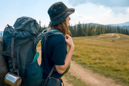 A young girl in a hat and with a backpack goes on an adventure, travels and enjoys nature, a woman looks at a picturesque view with meadows, forest and mountains on a sunny day.