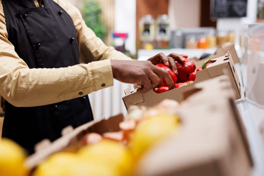 A modern supermarket owner arranges eco friendly products on a shelf. Selective focus on red peppers being organized by an african american person, highlighting local and sustainable options.