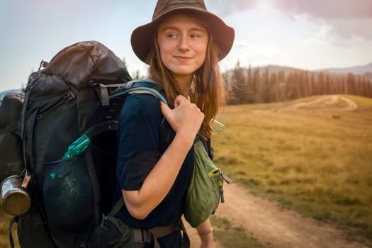 Young beautiful girl in a hat and with a tourist backpack walks through the fields, meadows, hiking mountainous terrain with a forest on a sunny day. A woman travels, goes on an adventure in the wild.
