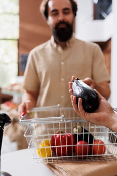 Close-up of bearded caucasian man giving natural eggplant to salesman for weighing. Male customer at cashier desk to check the weight of freshly harvested fruits and vegetables.