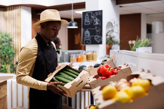 Owners arrange sustainable produce on wooden shelves where customers can pick freshly harvested items. Busy eco-store offers reusable packaging. Black man with an apron carrying a box of cucumbers,