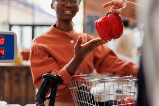 Close-up of black woman giving fresh red bell pepper to shopkeeper for weighing. African american client at cashier desk to check the weight of locally grown fruits and vegetables.