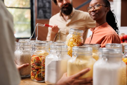 Close-up of african american girlfriend grasping a glass jar full of pasta while standing near caucasian boyfriend with basket. Multiethnic customers shop sustainably for healthy way of life.