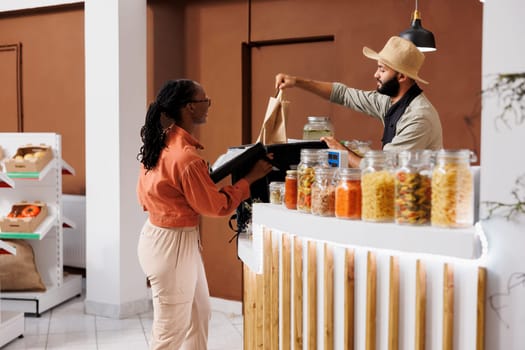 Merchant places a food order for delivery while packing homegrown organic vegetables in her backpack. African American courier arrives to collect bio supermarket products for local customers.