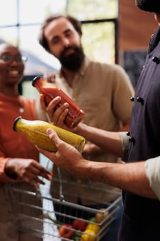 Happy boyfriend and girlfriend listening to a male vendor talk about different pasta sauces. Close-up of salesman holding jars of dressings to show to multicultural customers.