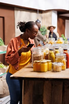 African American female customer compares prices, using her smartphone in a modern supermarket. Black lady inspecting things in glass containers at a bio-food store while holding a mobile device.