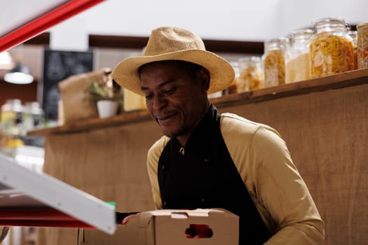 Detailed image shows a black man wearing an apron organizing a variety of newly harvested veggies on shelves. Close-up shot of an african american male vendor holding boxes of fresh, organic produce.