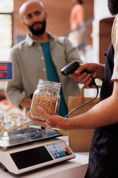 Bearded man waiting by the checkout counter while vendor scans the plastic free container having organic food product. Salesperson checking the price of the glass jar filled with natural soybeans.