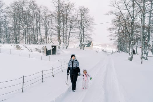 Mother and a little girl walk holding hands along a snowy road in a village on the edge of a forest. High quality photo