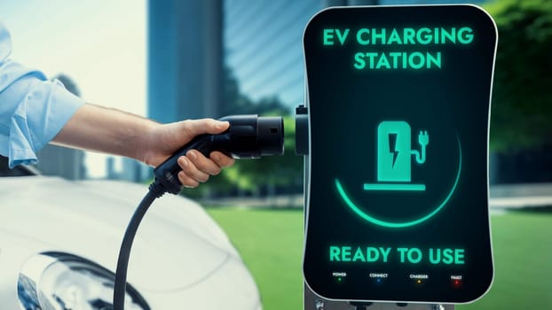 Businessman pull EV charger to recharge his electric car's battery from charging station in green eco city park background. Future innovative EV car and energy sustainability. Peruse