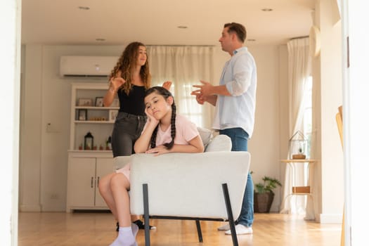 Annoyed and unhappy young girl sitting on sofa trapped in middle of tension by her parent argument in living room. Unhealthy domestic lifestyle and traumatic childhood develop to depression.Synchronos