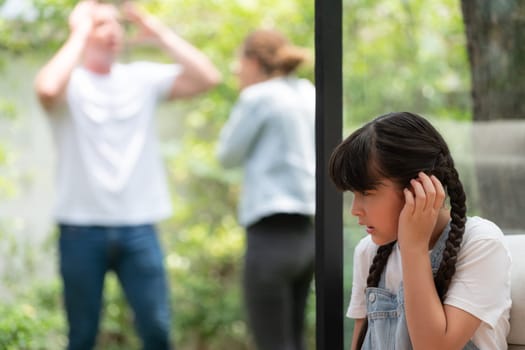 Stressed and unhappy young girl huddle in corner, cover her ears blocking sound of her parent arguing in background. Domestic violence at home and traumatic childhood develop to depression. Synchronos