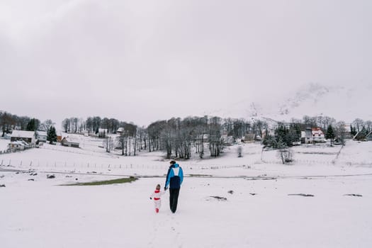 Father and a small child walk holding hands through a snowy pasture towards the village. Back view. High quality photo