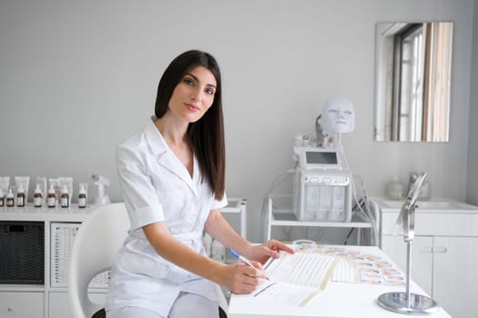 Portrait of confident dermatologist examining patient report at desk. Happy female cosmetologist working in modern medical office. Woman in white uniform sitting near modern equipment.