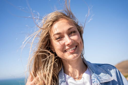 Closeup portrait of happy smiling beautiful woman with blond windswept hair. Cheerful female tourist on sea shore against blue sky. Lady enjoys summer vacation.