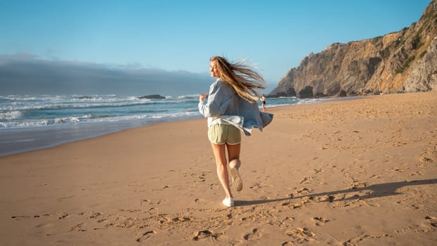 Rear view of smiling young woman running on beach. Happy female in denim shirt and shors looking back at camera along sea shore. Blond lady enjoying vacation against blue sky and mountain.