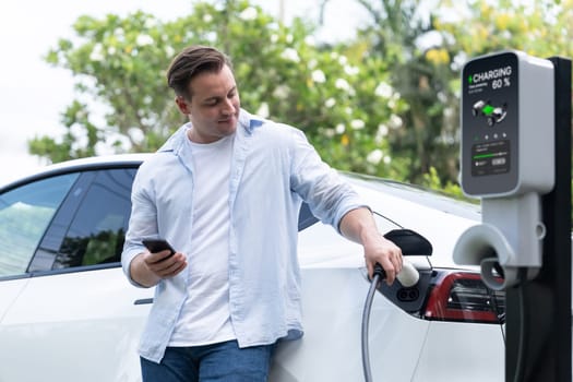 Modern eco-friendly man recharging electric vehicle from EV charging station, using Innovative EV technology utilization for tracking energy usage to optimize battery charging on smartphone.Synchronos